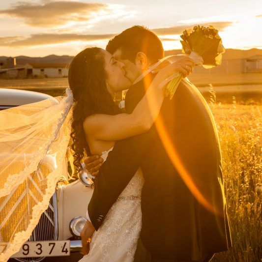 Novios abrazados con coche histórico a la puesta del sol