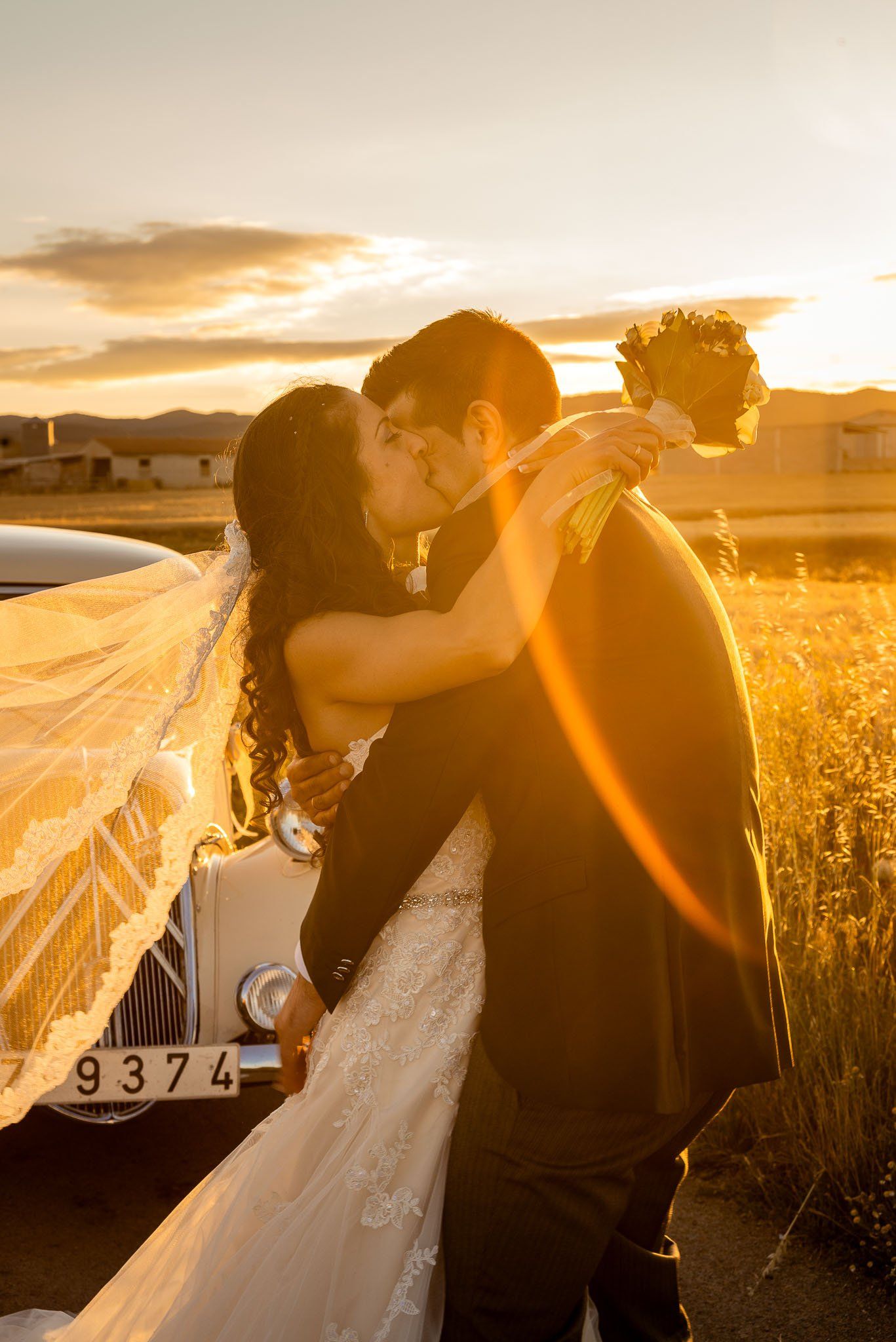 Novios abrazados con coche histórico a la puesta del sol